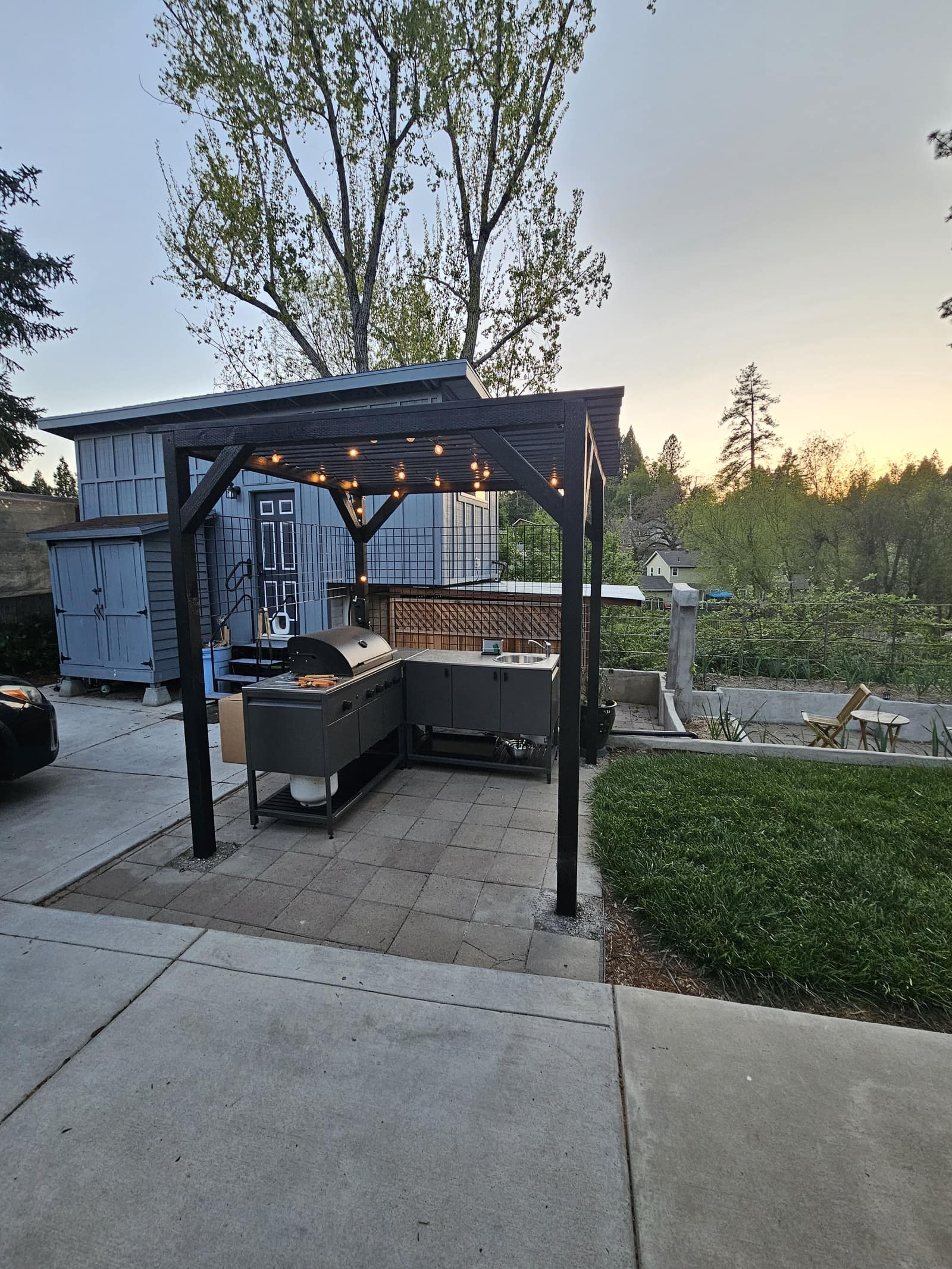 Outdoor kitchen under a timber pergola, with string lights and the garden beyond.