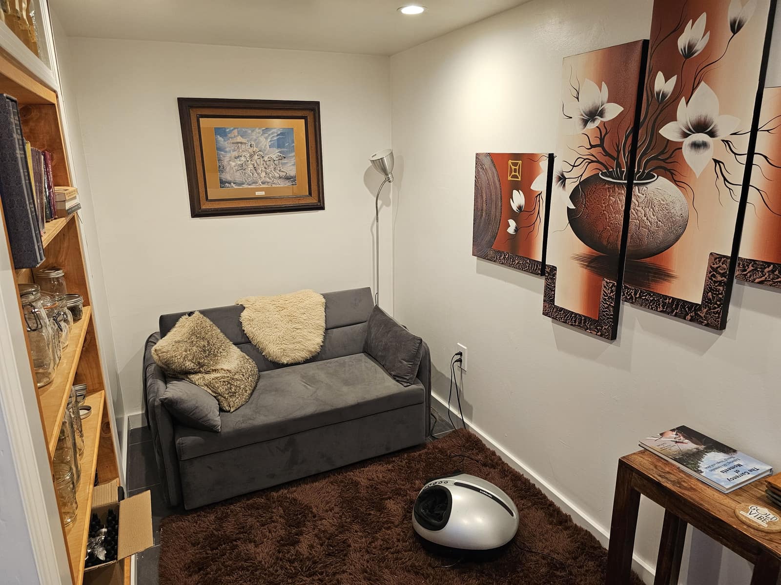 Den in Unit B with a gray sofa, wood shelves, and a large floral painting.